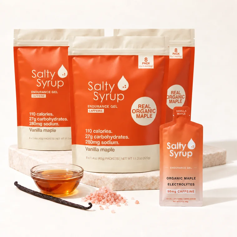 Three identical Salty Syrup Caffeinated bags on a stone block, each with a small 8 PACK badge, and one gel pouch in front, beside maple syrup, a vanilla pod and pink salt. Nothing in the frame says 24.