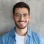 A smiling man in glasses and a denim shirt, standing against a plain background.