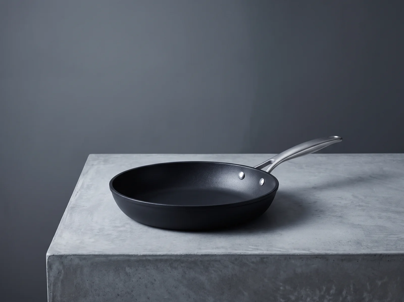 A black nonstick frying pan with a riveted stainless-steel handle, resting on a grey concrete surface.