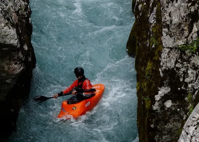 A person in an orange kayak paddling down a rough, narrow river.